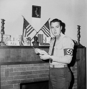 Ralph Forbes, 24, self styled western division commander of the American Nazi Party, poses beside the fireplace in his house in Glendale, Ca., Dec. 27, 1964, which the Party has leased as its western headquarters. Notice the picture of Hitler above the crossed American flags. Meanwhile city officials, clubs and churches are fuming because the Nazi group has located in its city. (AP Photo/David F. Smith)